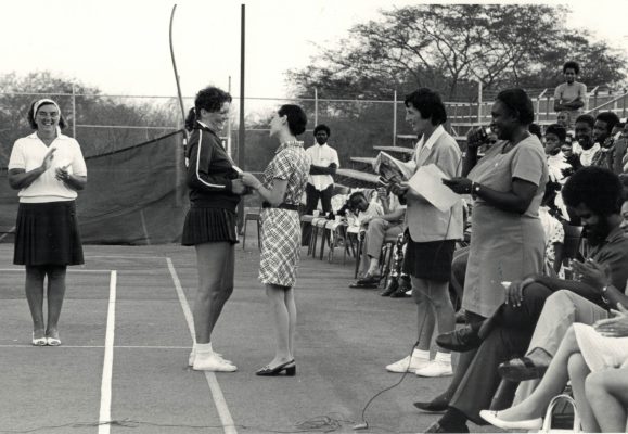 Linda Such receiving a souvenir spoon from Miss George Wyatt, wife of Managing Director of Jamaica Milk Products Ltd, manufacturers of Milo, main sponsor to the tour by England.  To the left is England Manager/Coach Mrs Mary French.  Also standing are (left to right) Miss Jo Wigman, Manager/Coach Jamaica National Squad and Miss Leila Robinson.  Seated (left to right) Senator Paul Miller, Parliamentary Secretary in the Ministry of Youth and Community Development and Mrs Dorothea Morris, Honorary Secretary Jamaica Netball Association. | Astley Chin, Photographer